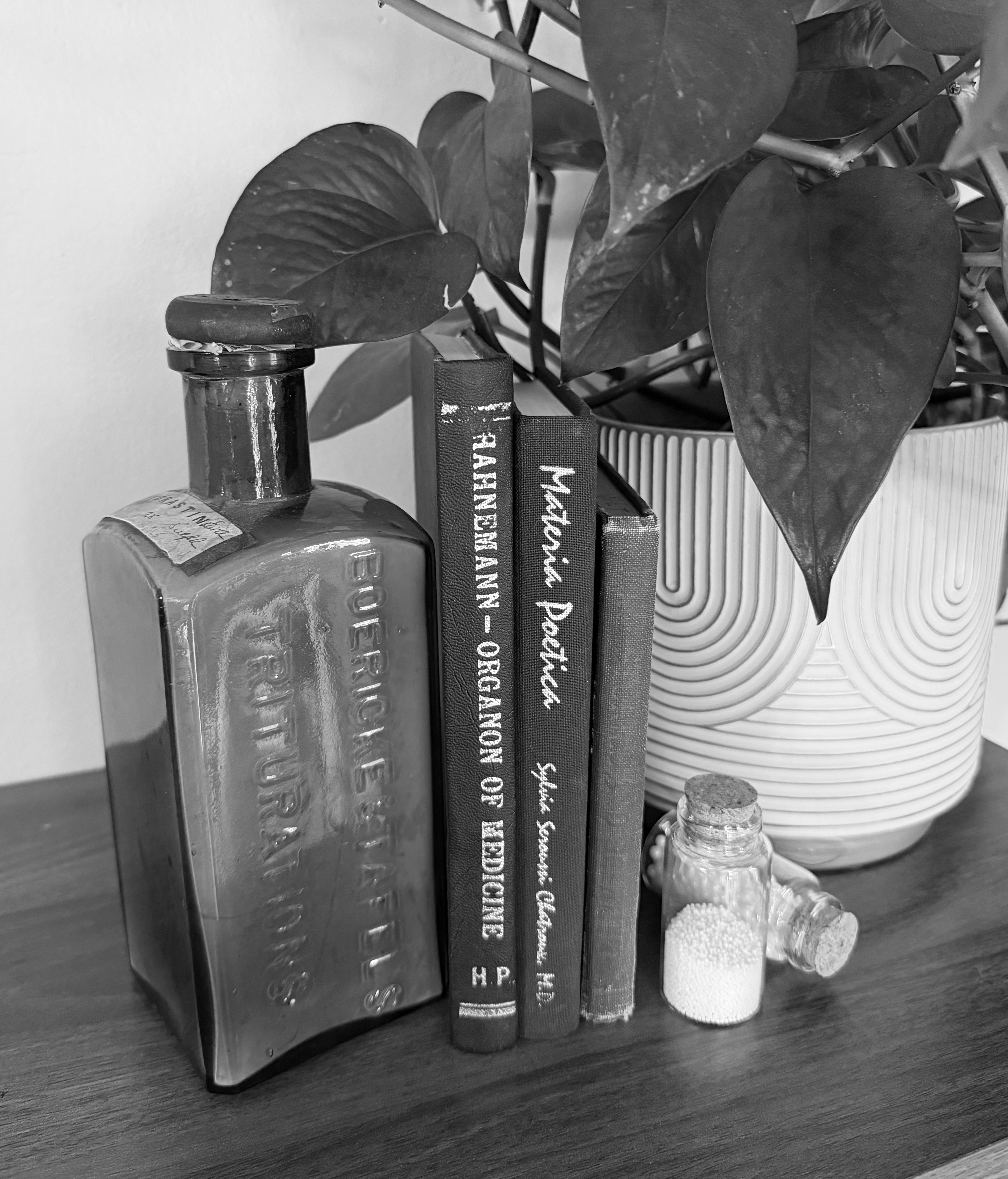 Black-and-white still life with antique Boericke and Tafel pharmacy bottles, homeopathic books, mortar and pestle, candle, and remedy vial